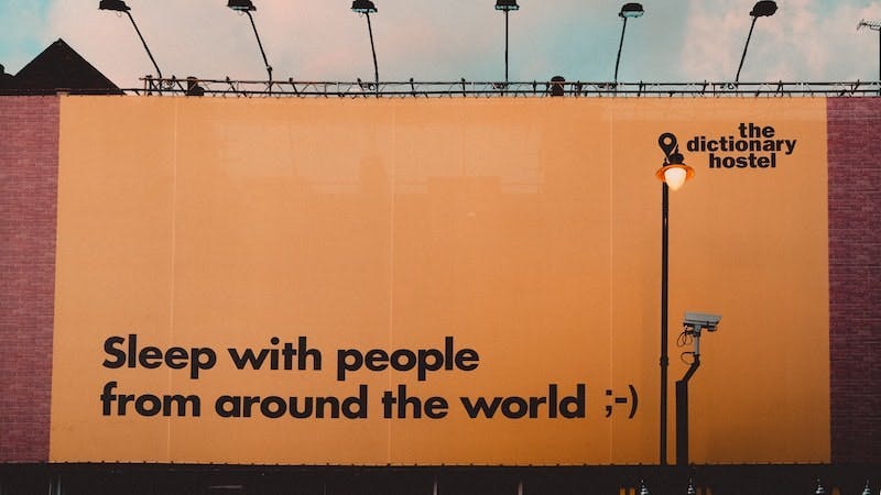 A billboard promoting the Dictionary hostel, the phrase “Sleep with people from around the world” is displayed